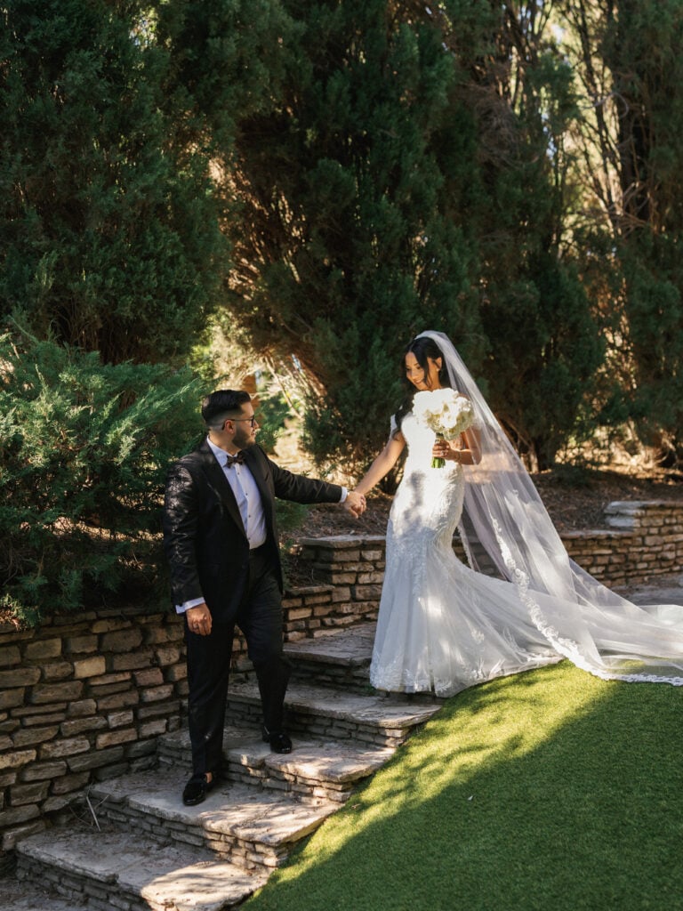 Elegant bride in a lace mermaid wedding dress holding a white peony bouquet walks down stone steps with the groom during their destination wedding travel at a California estate.