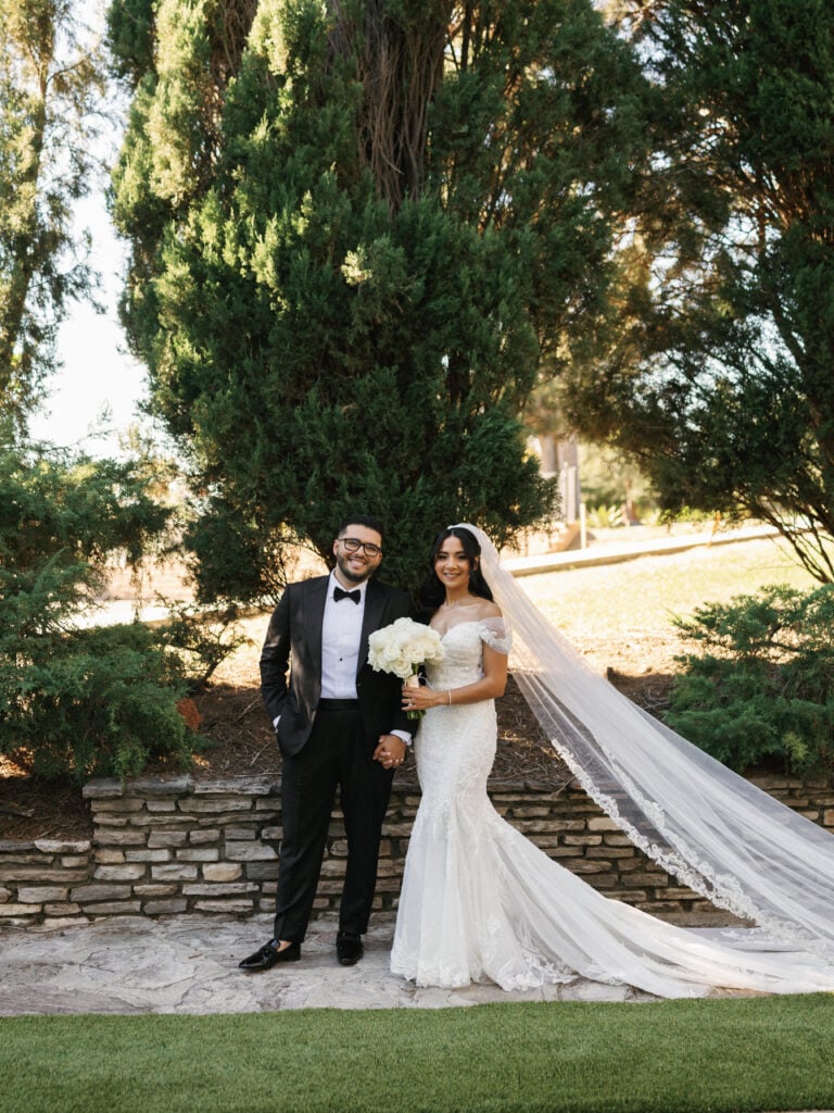 Smiling bride and groom holding hands by a stone wall under tall trees during their destination wedding travel at a scenic California estate.
