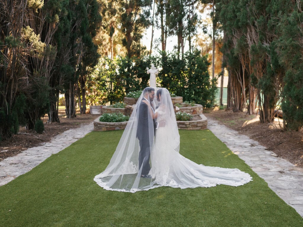 Romantic bride and groom embracing under a long lace-trimmed veil on a green lawn during their luxury destination wedding travel at a California estate.