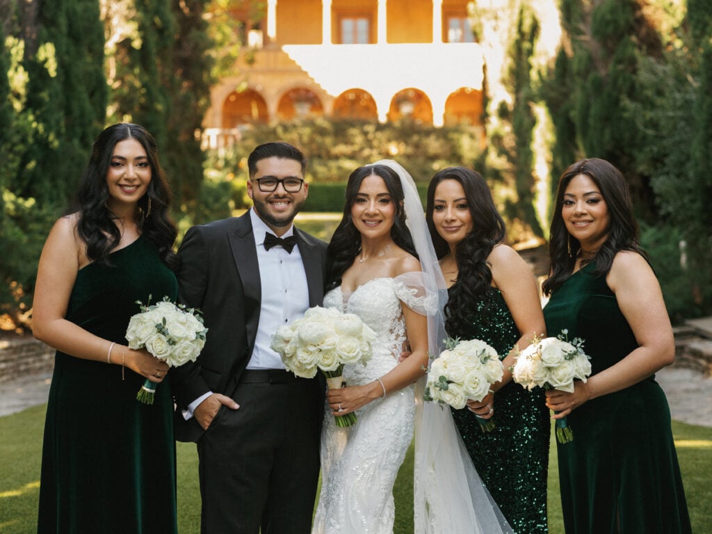 Elegant bride and groom posing with bridesmaids in emerald green velvet dresses holding white rose bouquets during their destination wedding travel celebration.