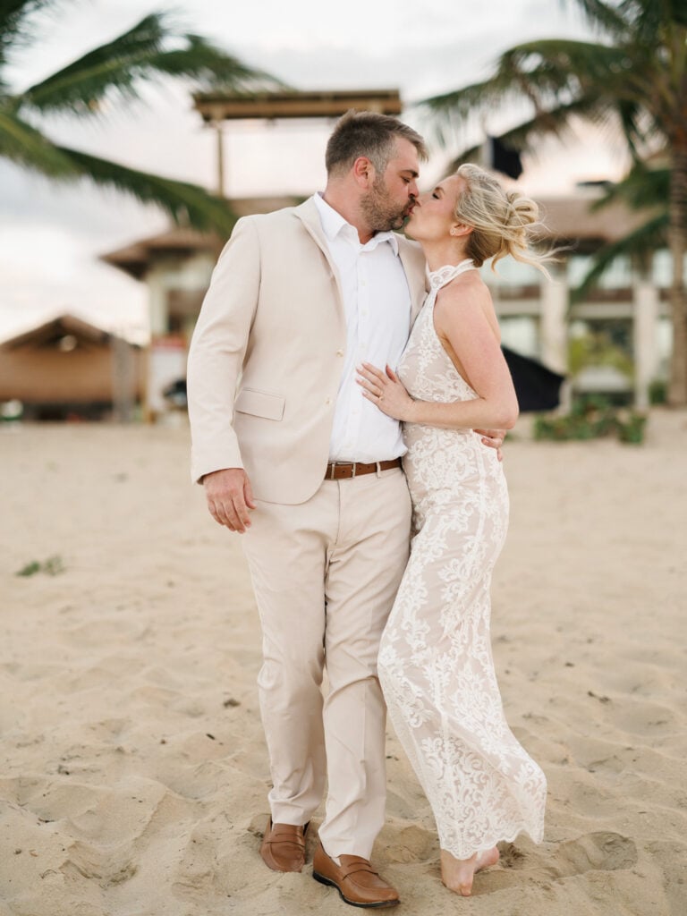 Romantic bride in a lace halter neck dress and a groom in a beige linen suit kissing on a sandy beach during their wedding weekend coverage.