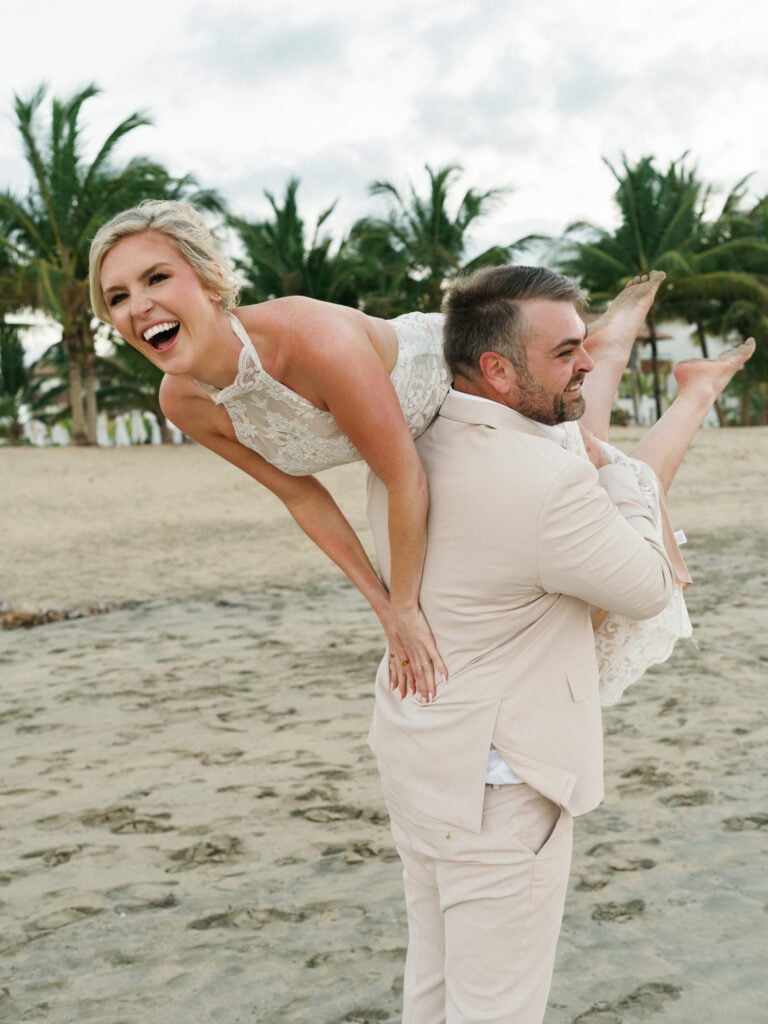Joyful groom carrying his laughing bride on a tropical beach with palm trees, capturing a fun candid moment for editorial wedding weekend coverage.