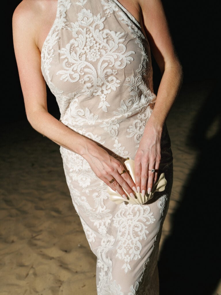 Close-up of a chic lace halter neck wedding dress paired with a champagne seashell clutch bag, inspiring high-end style for wedding weekend coverage.