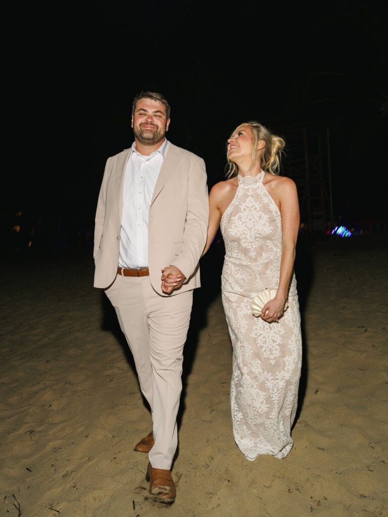 Smiling bride in a lace halter neck reception dress walking hand-in-hand with her groom in a beige suit on the sand during their wedding weekend coverage.