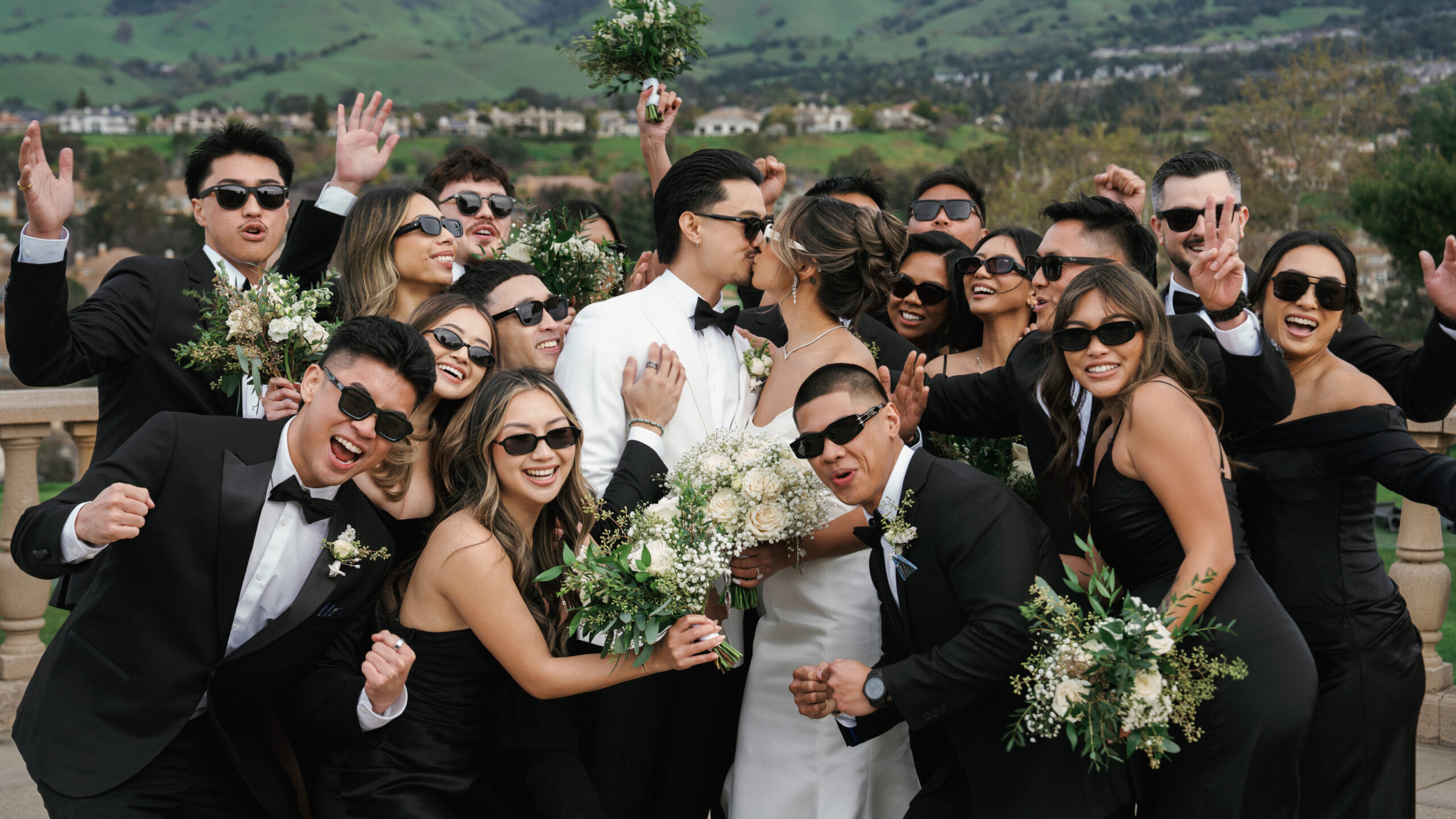 Newlyweds kiss among a wedding party wearing sunglasses outdoors.