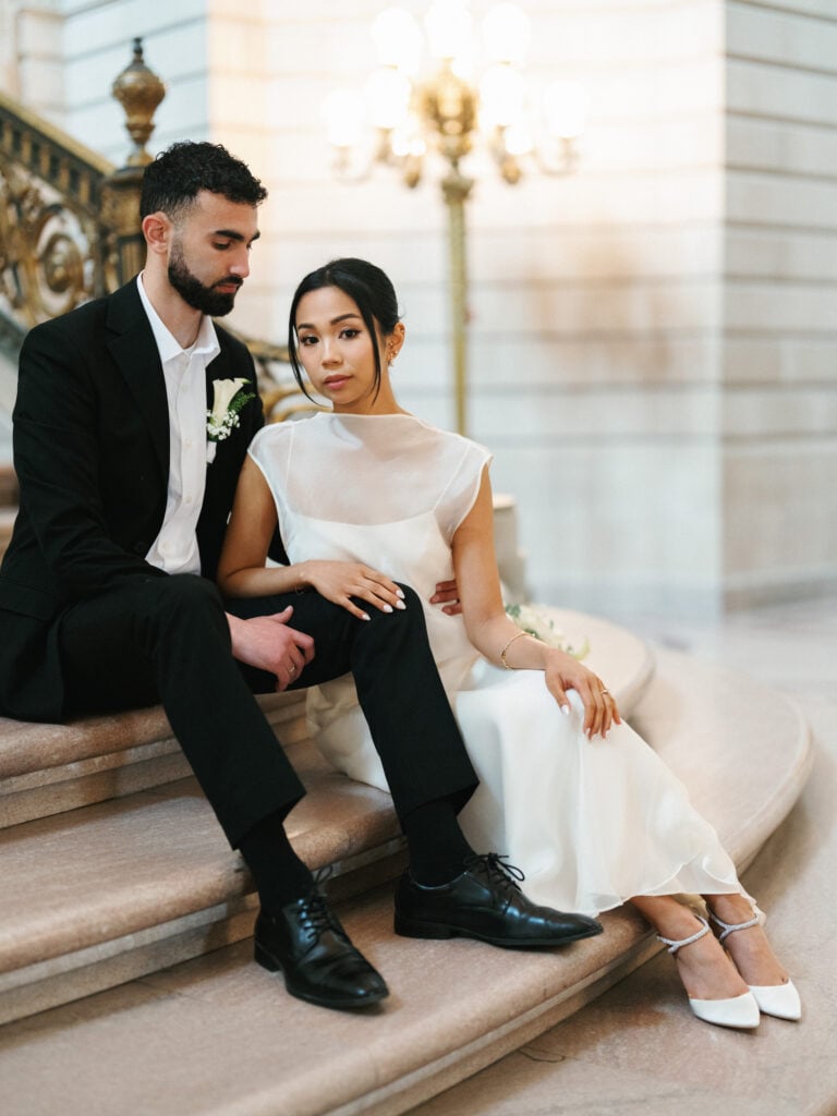 Elegant bride in a high-neck wedding dress with ankle-strap heels sitting on stone steps with her groom, inspiring editorial wedding photography at San Francisco City Hall.