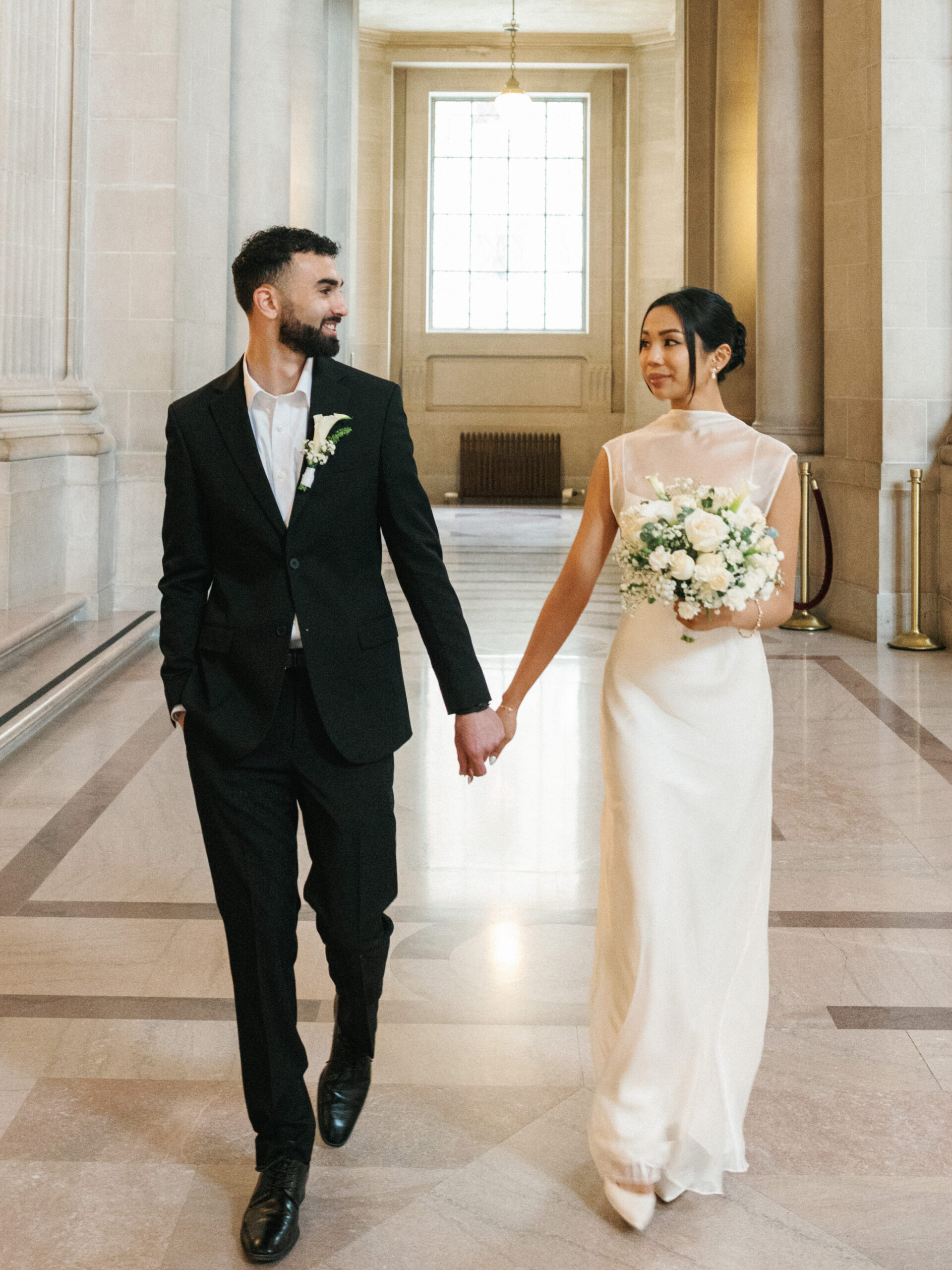Editorial wedding photo ideas featuring a modern couple walking hand-in-hand through the grand marble halls of San Francisco City Hall.