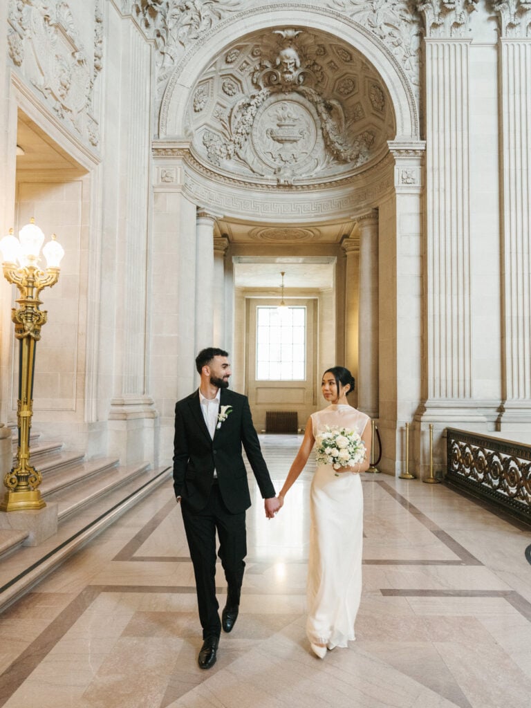 Candid bride and groom holding hands while walking through the grand Beaux-Arts interior of San Francisco City Hall, illustrating fluid editorial wedding photography.