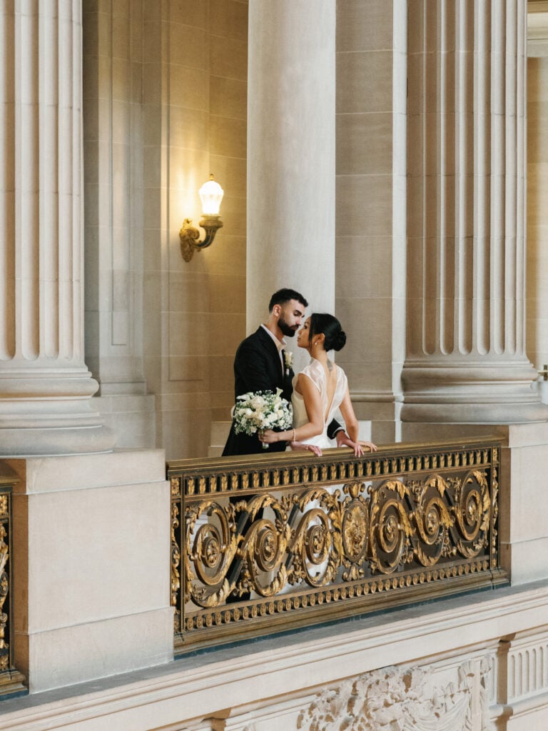 Elegant bride in a high-neck wedding dress embracing her groom by an ornate gold balustrade near massive limestone columns during an editorial wedding photography shoot.