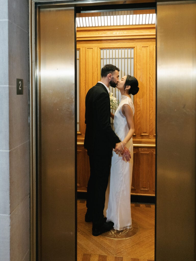 Candid bride and groom sharing an intimate kiss inside a historic wood-paneled elevator at San Francisco City Hall, capturing the fluid essence of editorial wedding photography.