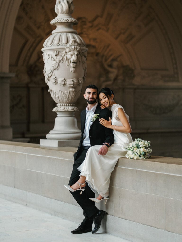 Relaxed bride in a sleek high-neck wedding gown sitting with her groom next to an ornate stone urn, posing naturally for high-end editorial wedding photography.
