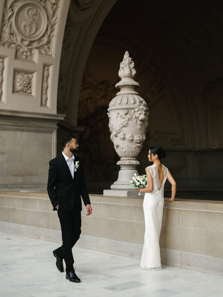 Candid bride in a minimalist open-back gown and groom looking at each other near a historic stone urn at San Francisco City Hall during editorial wedding photography.