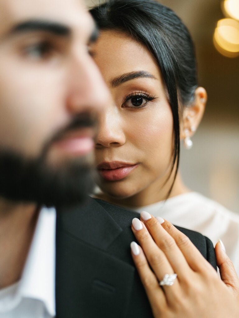 Close-up of a bride with an elegant updo leaning on her groom's shoulder, showcasing her diamond engagement ring during an editorial wedding photography shoot.