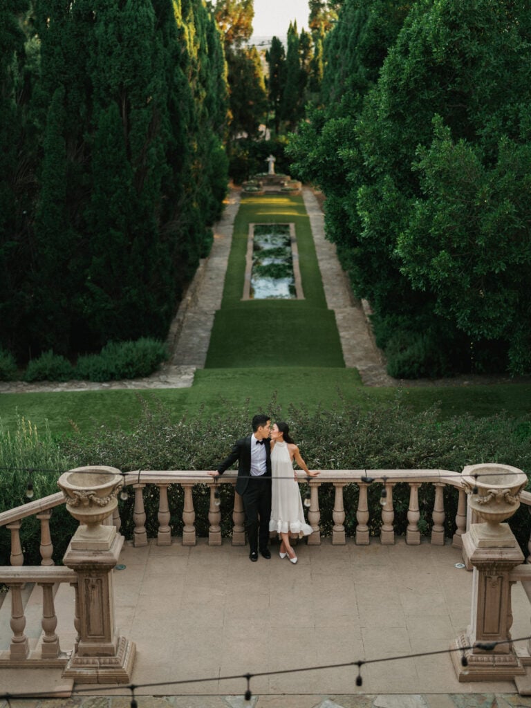 Chic couple kissing on an ornate stone balcony overlooking a dramatic cypress tree alley during their destination wedding travel at an Italian-style California estate.