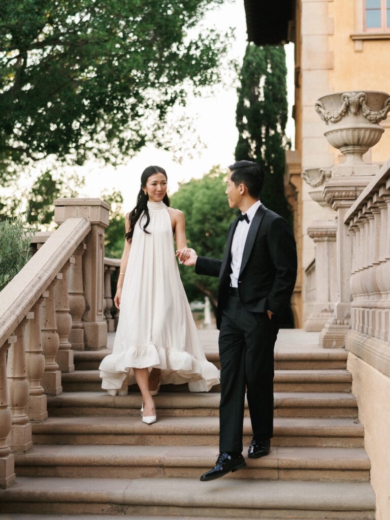 Elegant bride in a high-neck halter wedding dress holding hands with the groom while walking down stone steps during their destination wedding travel at a luxury California villa.
