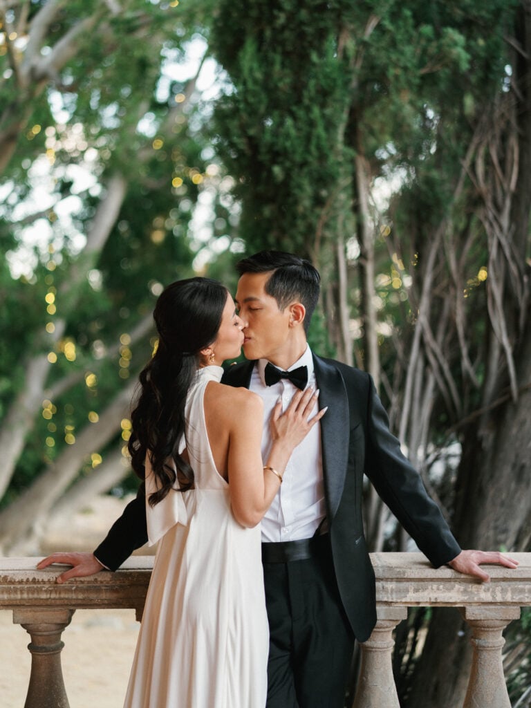 Chic bride and groom sharing a romantic kiss by a stone stone balustrade under lush green trees during their luxury destination wedding travel in California.
