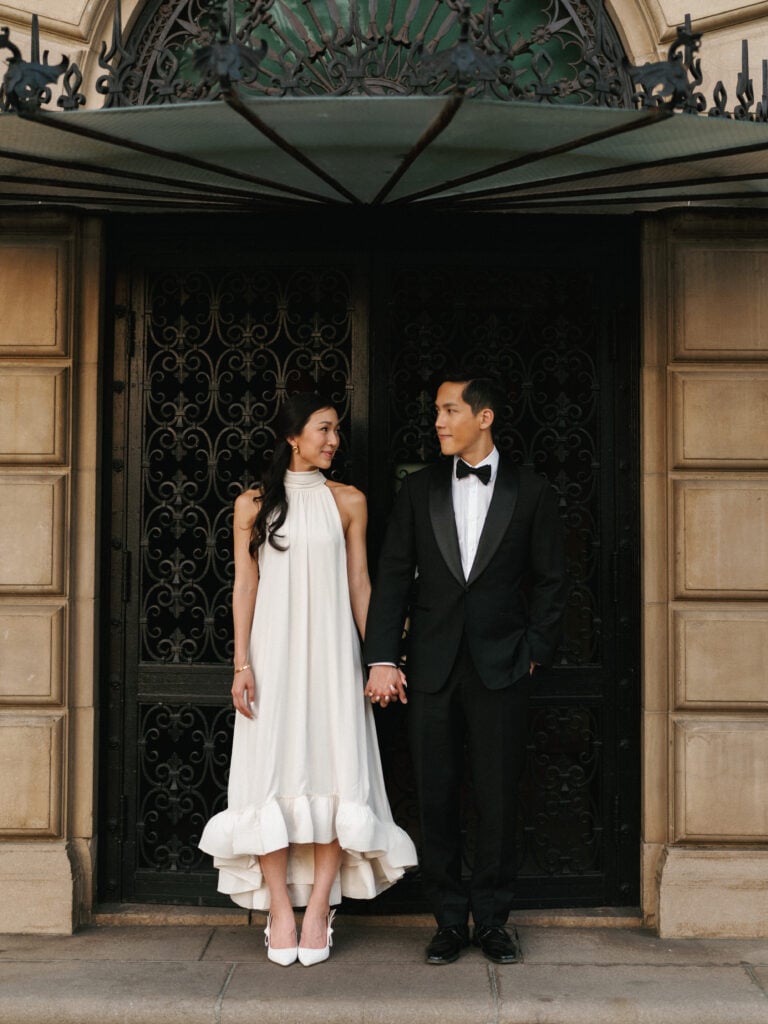Elegant bride in a high-neck halter wedding gown holding hands with her groom in a tuxedo before ornate wrought-iron doors during their destination wedding travel.