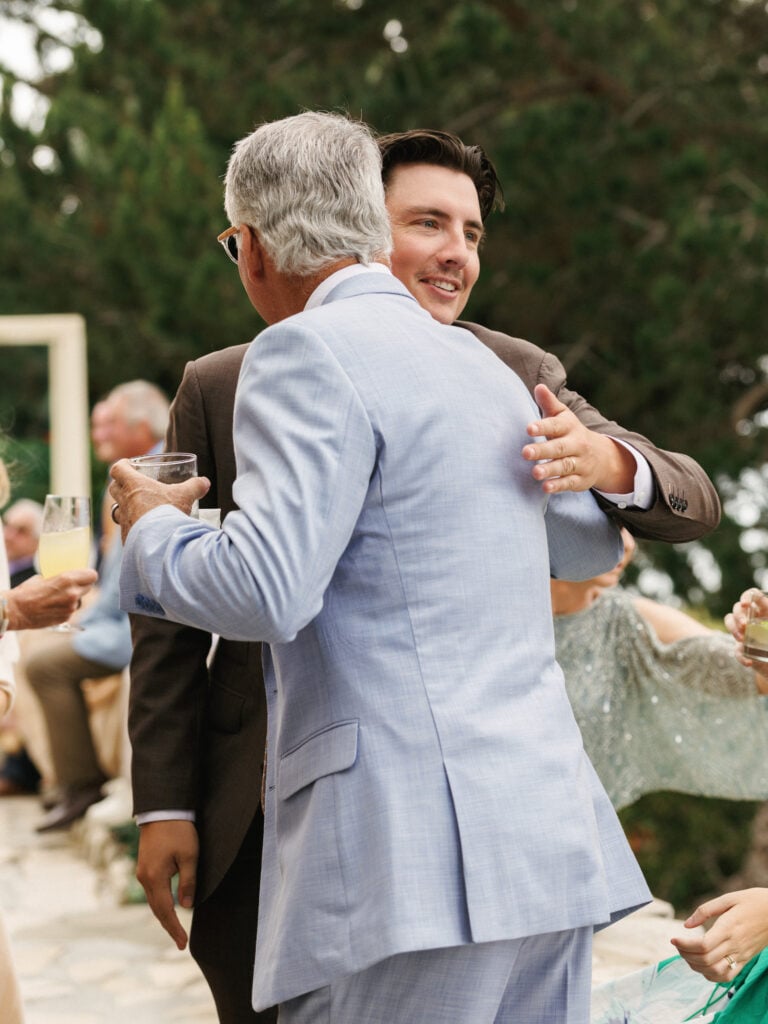 Groom in a brown suit embracing a wedding guest in a light blue summer suit during an outdoor cocktail hour included in wedding weekend coverage.