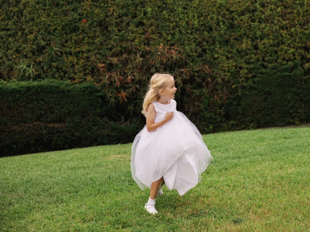 Cute flower girl in a fluffy white tulle dress running joyfully across a green lawn, capturing a sweet spontaneous moment of professional wedding weekend coverage.