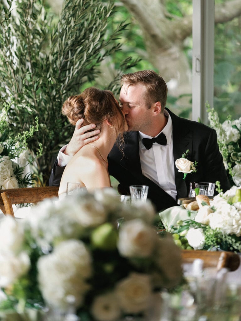 Romantic bride and groom sharing a candid kiss at a luxury head table adorned with lush white roses and olive greenery during their editorial wedding weekend coverage.