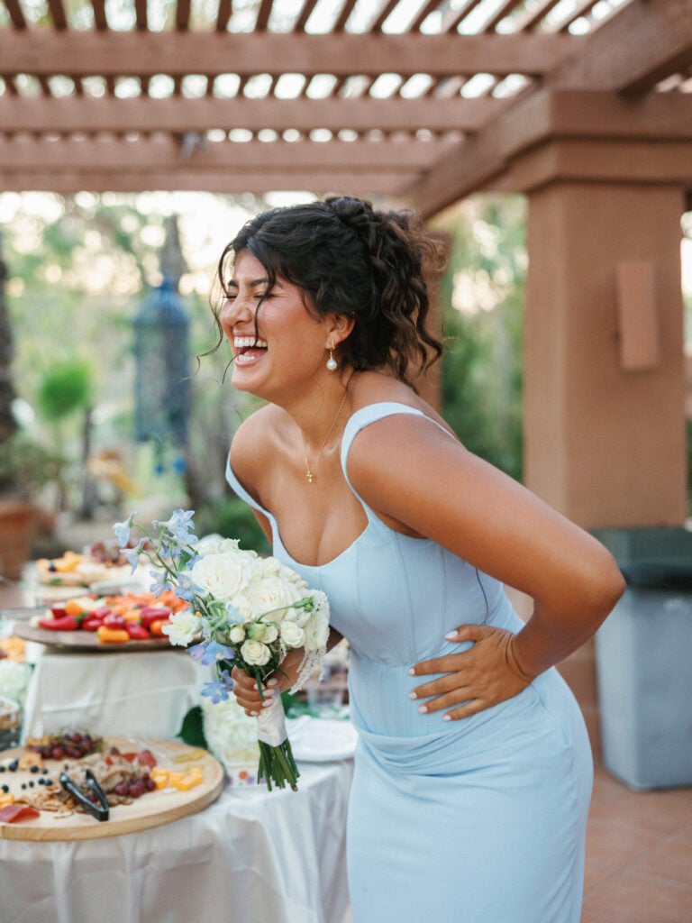 Laughing bridesmaid in a dusty blue corset dress holding a white and blue delphinium bouquet by a cocktail buffet table during a vibrant wedding weekend coverage event.