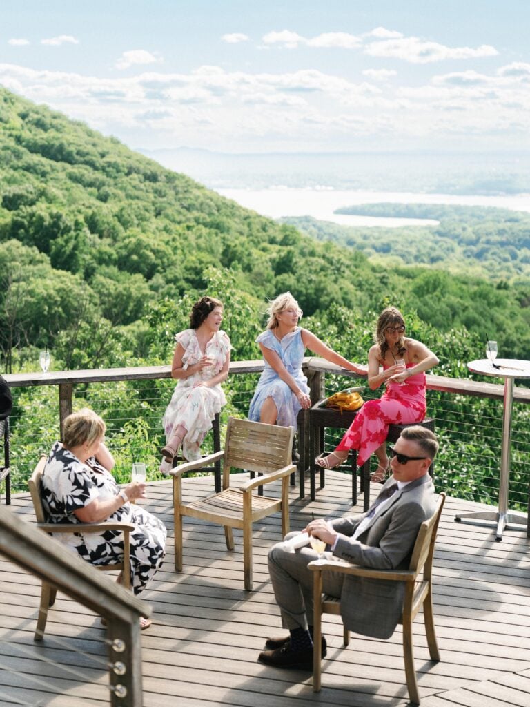 Wedding guests relaxing on a luxury wooden terrace with a panoramic view of lush green hills and a river valley during comprehensive wedding weekend coverage.