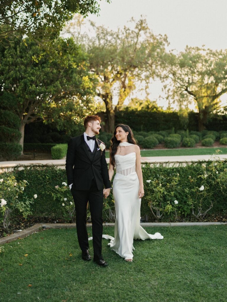 Elegant bride in a strapless corset bodice wedding dress with a chiffon scarf walking with her groom on a green lawn, showcasing modern editorial wedding photography.