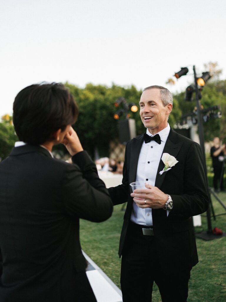 Sophisticated groom or guest in a classic black tuxedo laughing during an outdoor cocktail hour, capturing candid moments of wedding weekend coverage.
