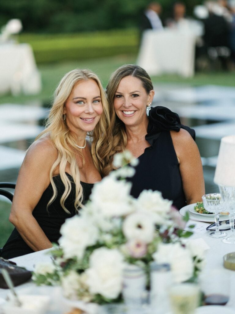 Elegant female wedding guests in chic black cocktail dresses smiling at a luxury outdoor reception table during editorial wedding weekend coverage.