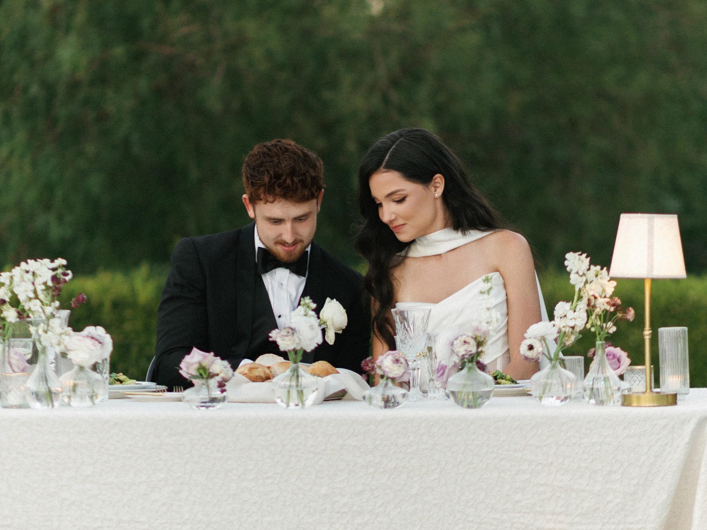 Elegant bride in a high-neck halter wedding gown and a groom in a classic tuxedo sitting at a luxury reception table with pastel florals and a small cordless lamp during their wedding weekend coverage.