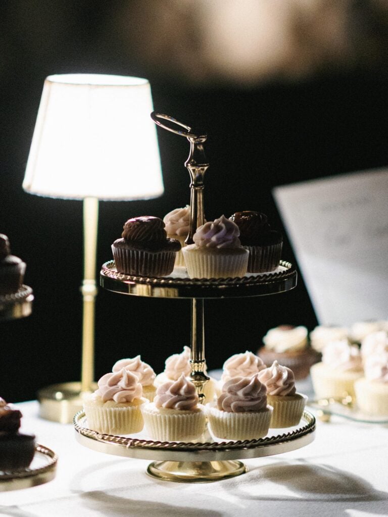 Elegant tiered dessert stand filled with pink and chocolate cupcakes next to a small table lamp at a night reception, inspiring detailed wedding weekend coverage ideas.