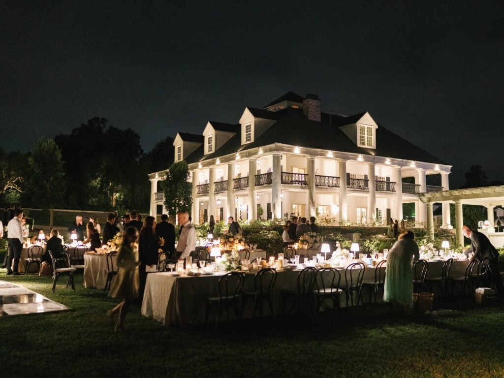 Luxury outdoor wedding reception illumination with long tables on a green lawn in front of a lit southern colonial mansion, captured during editorial wedding weekend coverage.