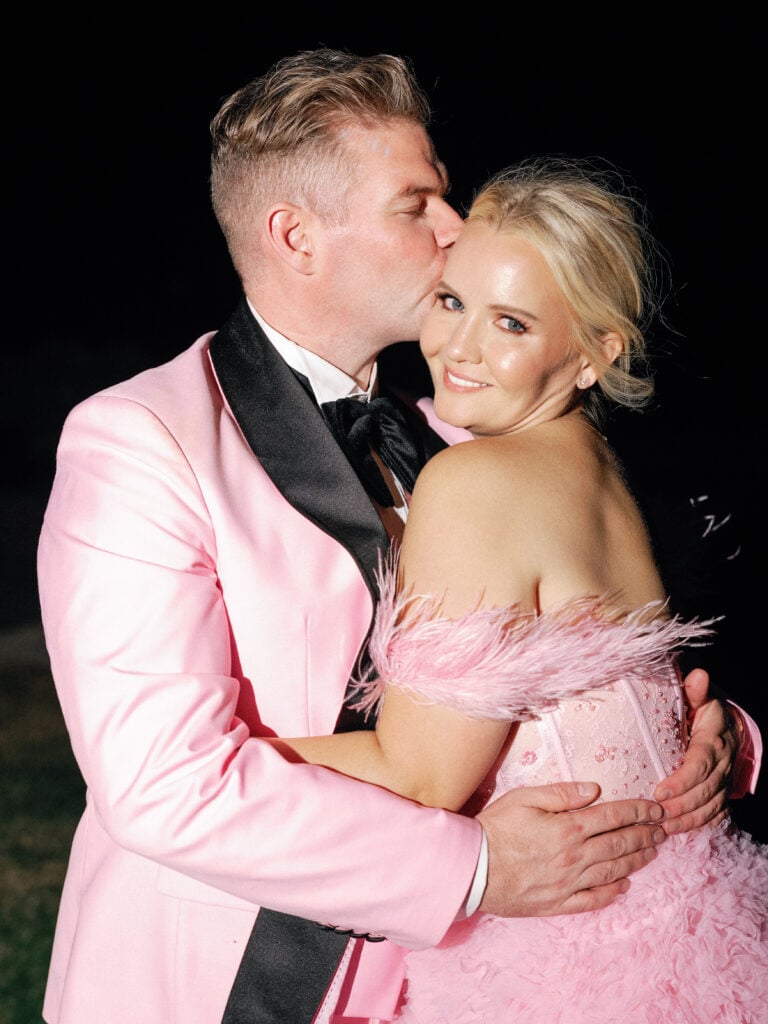 Romantic groom kissing his bride on the forehead during an outdoor night celebration, showcasing a matching pink tuxedo and pink tulle feather gown for creative editorial wedding photography.