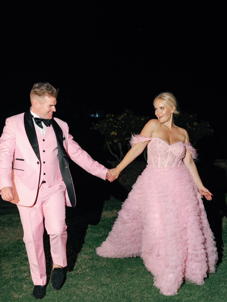 Bride in a pink ruffle gown and groom in a pink tuxedo running on a lawn at night, shot on film with flash by a Palm Springs wedding photographer.
