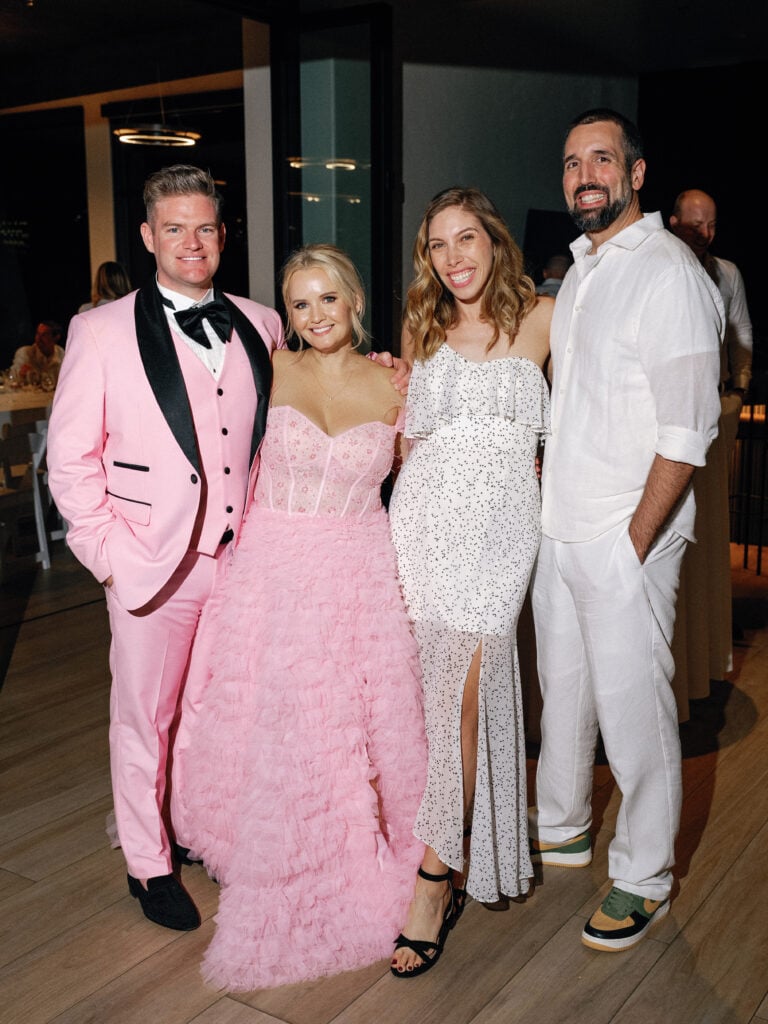 Chic couple in a matching pink tuxedo and ruffled tulle gown posing with stylishly dressed wedding guests during a vibrant wedding weekend coverage celebration.