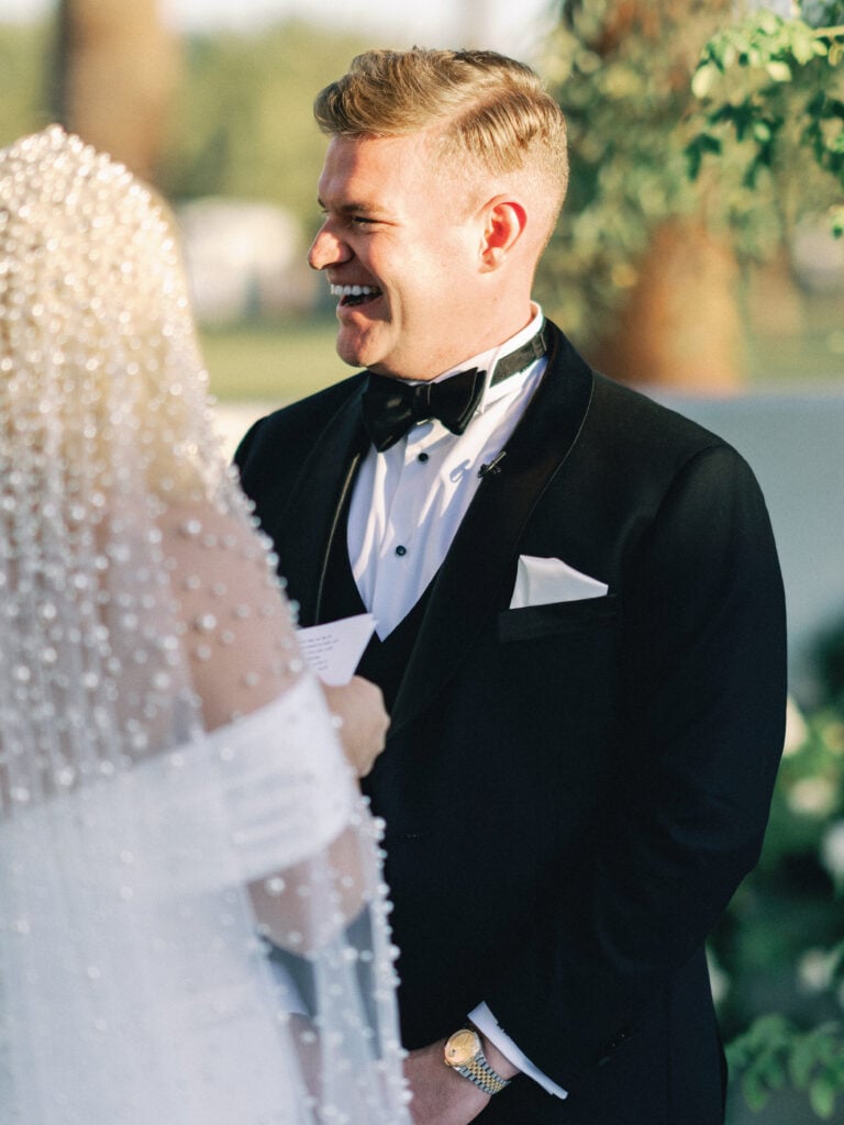 Groom in a black tuxedo smiling during wedding vows next to a bride wearing a pearl veil, shot on film by a Palm Springs wedding photographer.