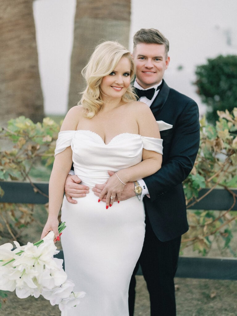 Bride holding a white lily bouquet while her groom embraces her from behind, captured on film by a Palm Springs wedding photographer.