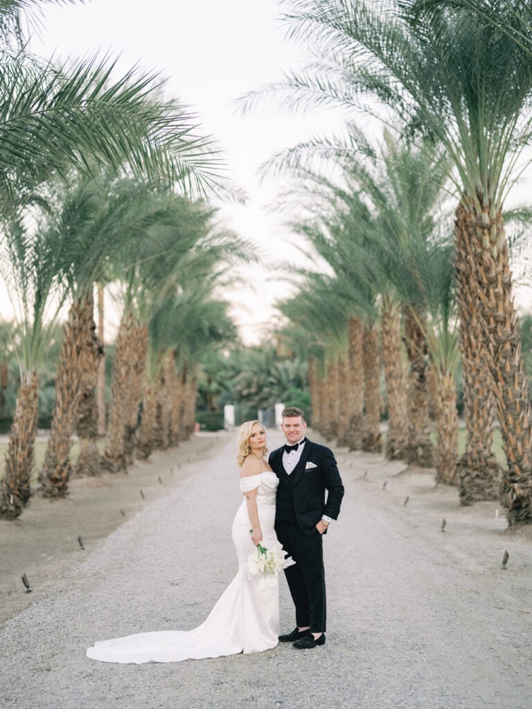Bride and groom posing on a path lined with tall palm trees, shot on film by a Palm Springs wedding photographer.