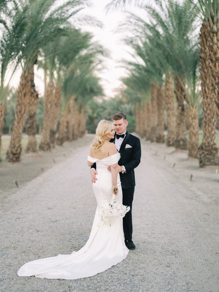 Elegant film wedding portrait of a couple embracing on a majestic palm tree avenue, curated by a professional Palm Springs wedding photographer.