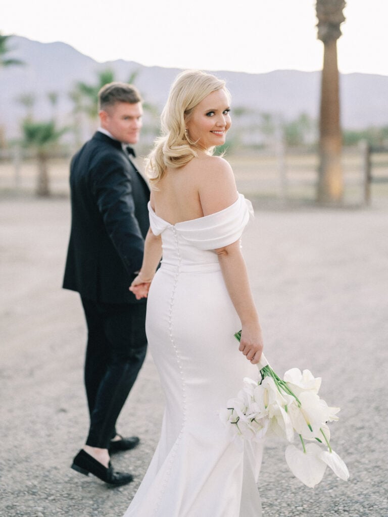 Candid film wedding photography of a smiling bride looking back while holding hands with her groom, captured by a Palm Springs wedding photographer.
