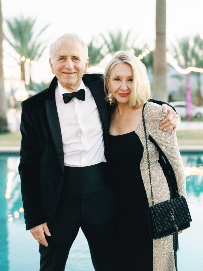 Parents of the bride posing by a pool in formal black tie attire with a luxury handbag, captured on film by a Palm Springs wedding photographer.