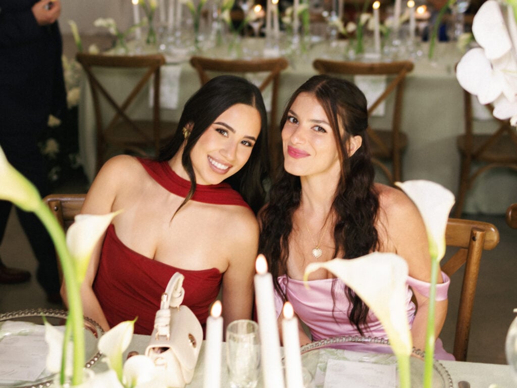 Beautiful bridesmaids in elegant burgundy and satin pink cocktail dresses smiling at a candlelit reception table during high-end wedding weekend coverage.