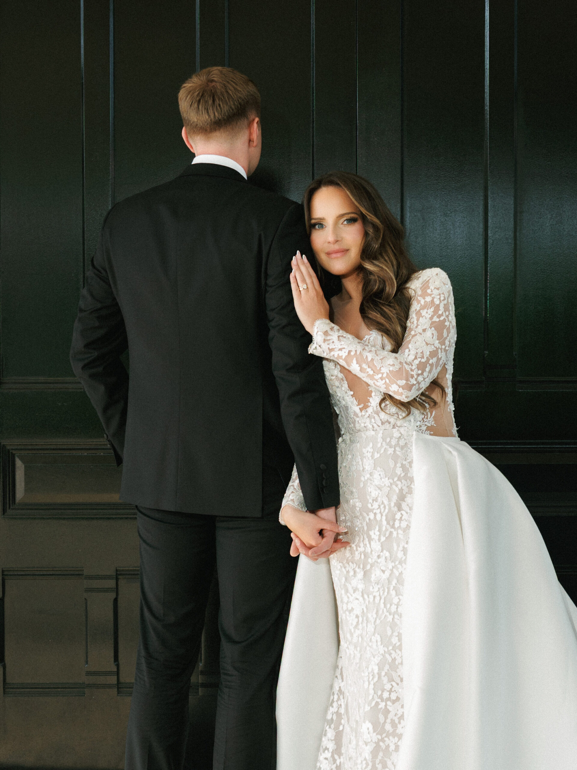 Bride in long-sleeve lace gown resting her head on groom's shoulder in front of dark wood paneling.