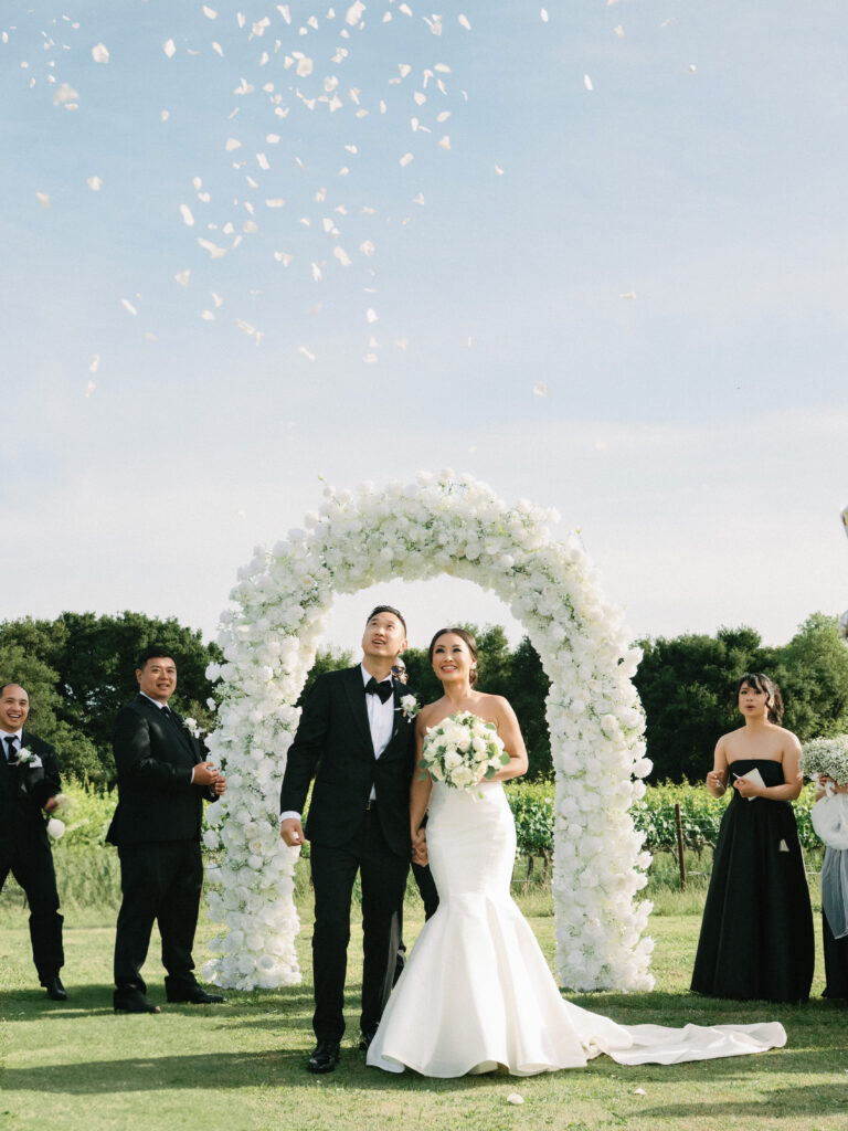 Joyful couple celebrating under a white flower arch during a wedding petal toss, shot on film by a Napa Valley wedding photographer.