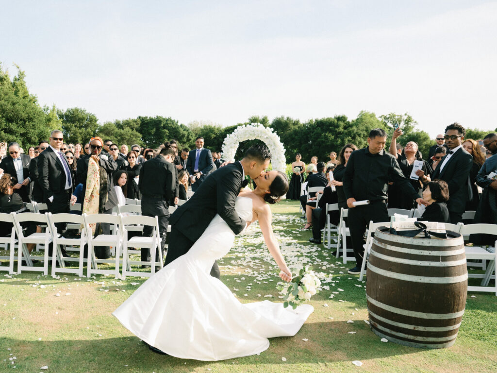 Editorial film wedding photo of a couple's first kiss at an outdoor altar with white chairs, flower arch, and rustic wine barrels, by a Napa Valley wedding photographer.