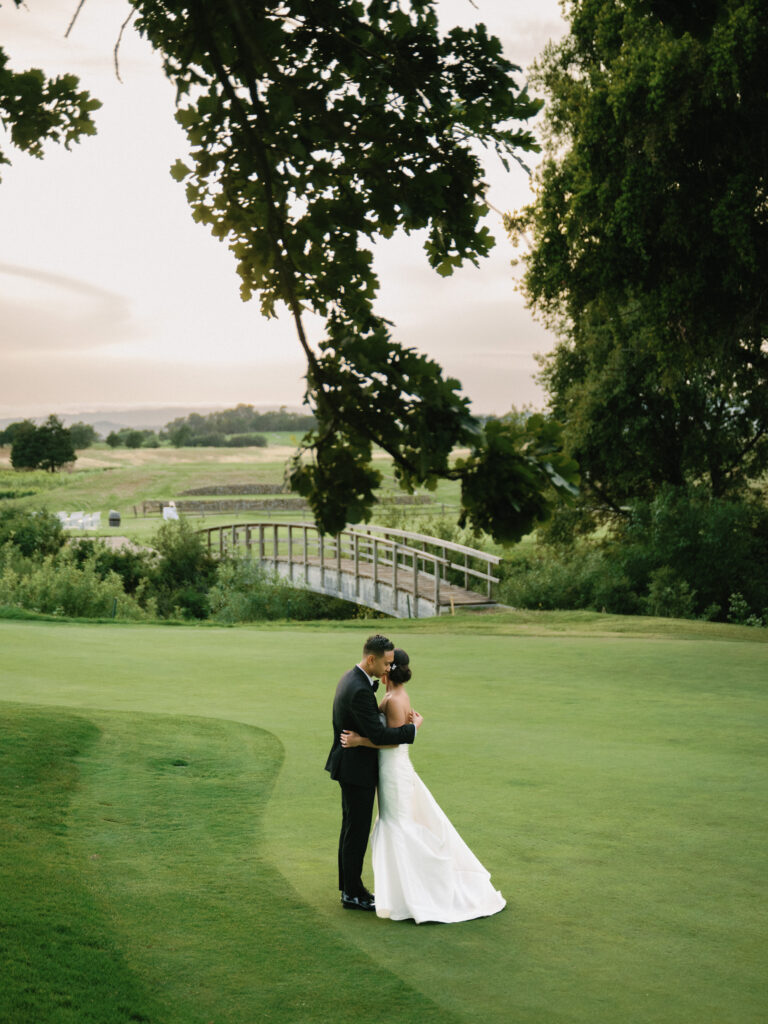 Wide-angle film wedding photography of a couple embracing on a vast green lawn with a rustic bridge and trees backdrop, taken by a Napa Valley wedding photographer.
