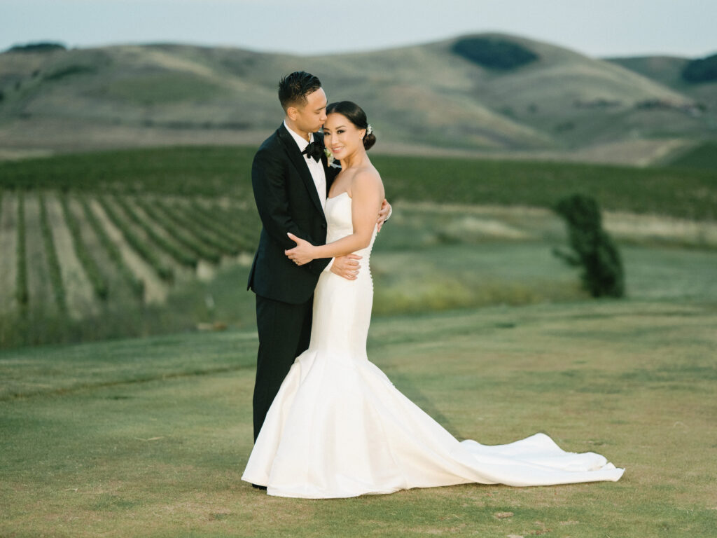 Elegant film wedding portrait of a couple embracing with lush Napa Valley vineyards and rolling hills in the background, curated by a professional wedding photographer.