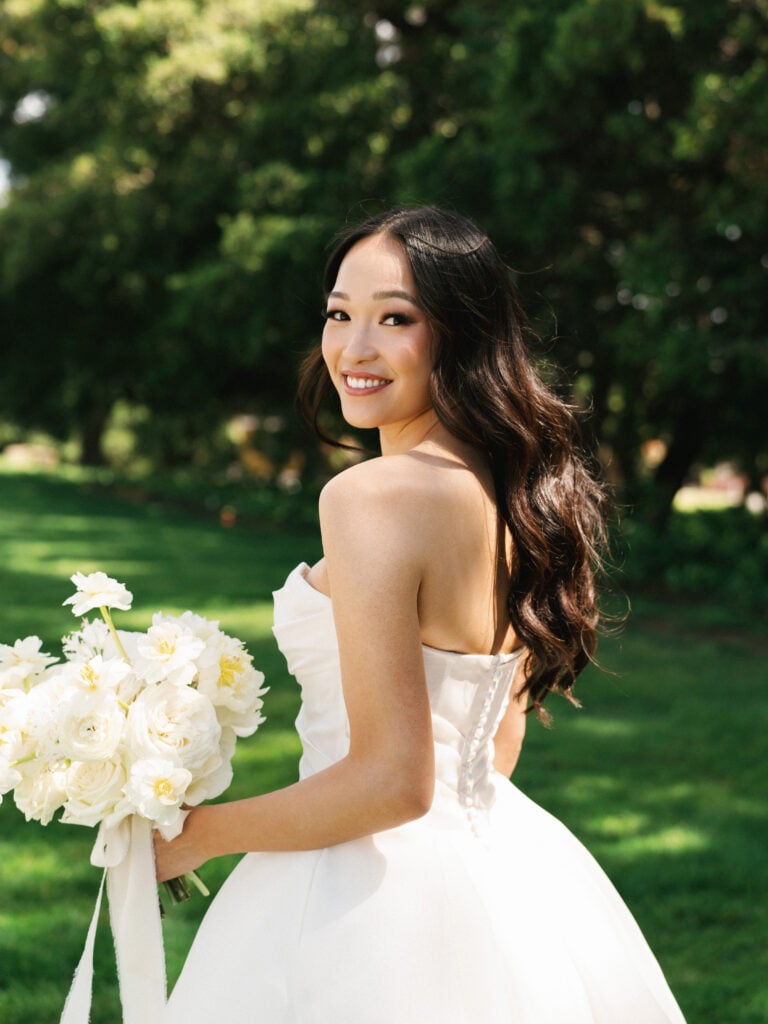 Smiling bride with long Hollywood waves looking over her shoulder while holding a white peony bouquet, showcasing relaxed editorial wedding photography.