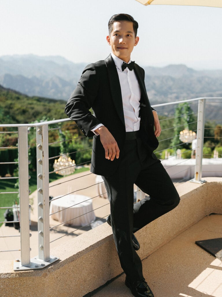 Groom in a classic black tuxedo and bow tie posing on a scenic mountain view terrace during a luxury destination wedding travel production in California.