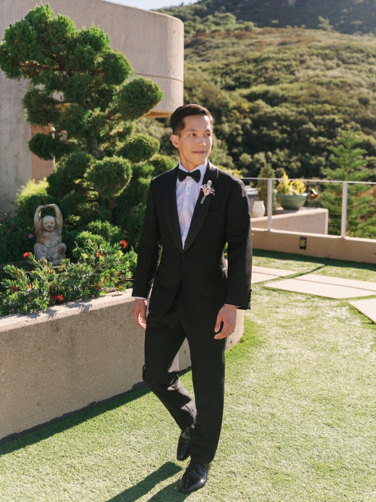 Groom in a tailored black tuxedo walking on a sun-drenched terrace overlooking the Malibu mountains during a luxury destination wedding travel production.